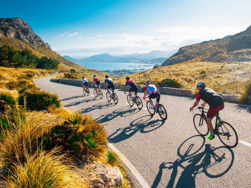 A group of road cyclists descending a hilly road with a view of the Costa Brava coastline.